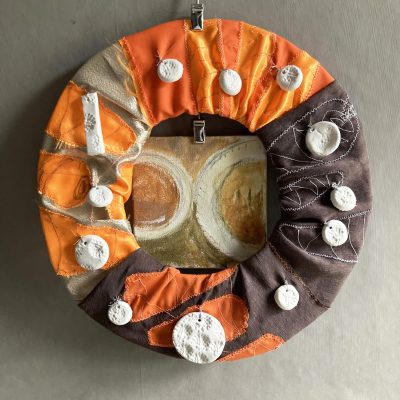 The Studio Snack wreath of leftovers: a hoop covered with cloth embroidered with fluorescent and luminous carrots, with clay acorn imprint beads sewn on and a small painting behind of an Eccles cake and a cup of coffee