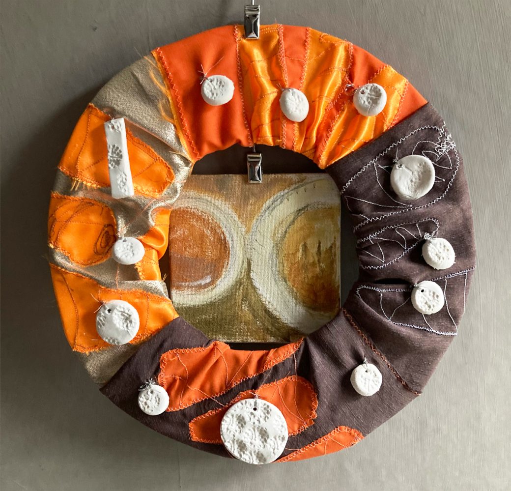 The Studio Snack wreath of leftovers: a hoop covered with cloth embroidered with fluorescent and luminous carrots, with clay acorn imprint beads sewn on and a small painting behind of an Eccles cake and a cup of coffee