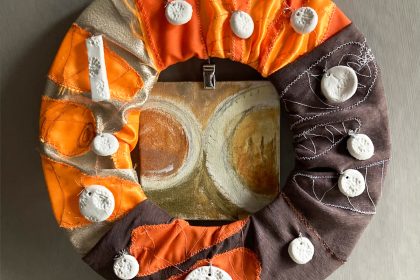 The Studio Snack wreath of leftovers: a hoop covered with cloth embroidered with fluorescent and luminous carrots, with clay acorn imprint beads sewn on and a small painting behind of an Eccles cake and a cup of coffee